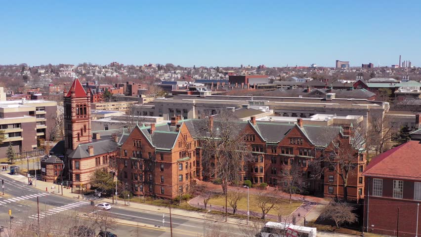 Aerial drone footage capturing expansive view of traditional red brick university campus buildings situated on corner of busy intersection in boston featuring bright sunny spring sky.