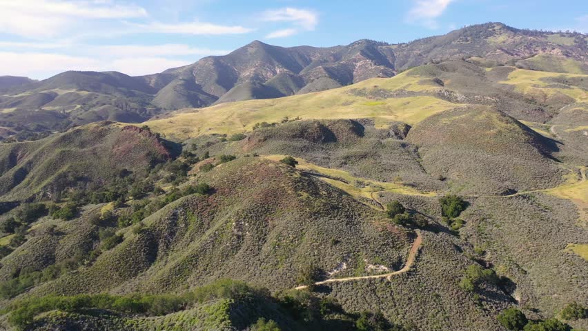 Aerial view captures beautiful rolling green hills and rugged mountain terrain in rural California. Sunlight highlights gentle slopes and textures of grassy landscape under blue sky.