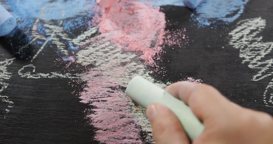 A hand is using a chalk piece to draw colorful patterns on a black surface. Various chalk colors are scattered around and the texture is visible.