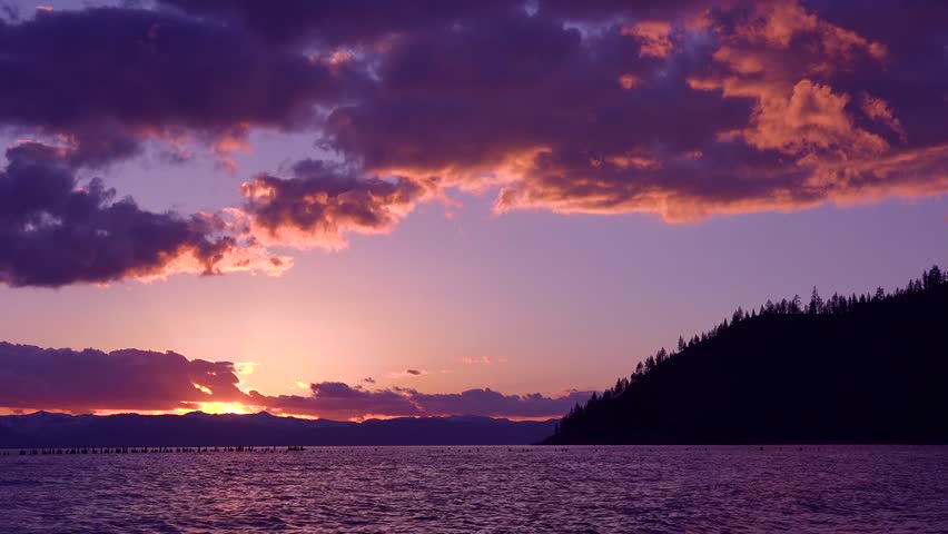 Breathtaking dusk landscape featuring vibrant violet clouds moving across sky. Peaceful water reflections at sundown with silhouetted trees. Cinematic mountain horizon and soft light.
