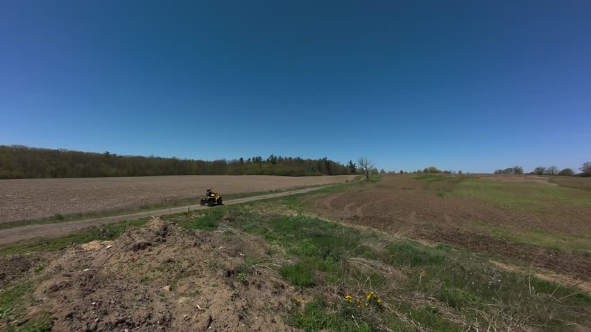 Drone tracking shot following an all-terrain vehicle riding along a dirt path between textured agricultural land and green grassy property on a sunny spring day in Milton Ontario.