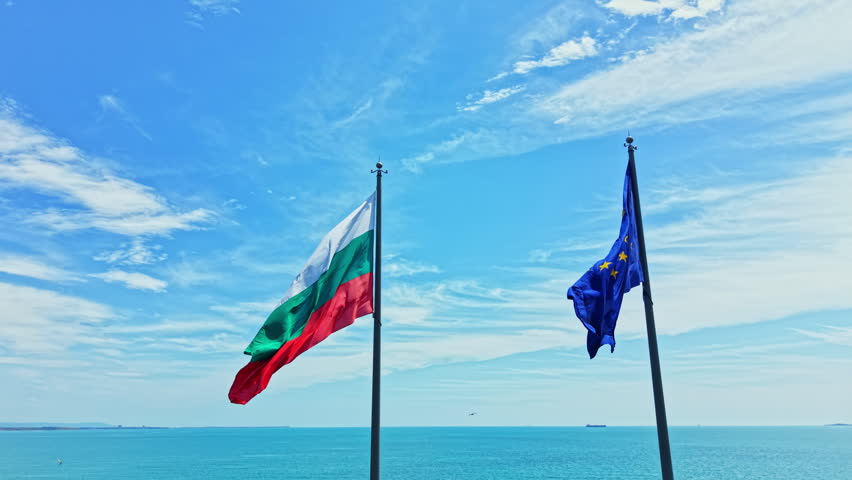 Flags of Bulgaria and the European Union fly next to each other above the ocean. The sky is mostly clear with some clouds, and the water appears calm.