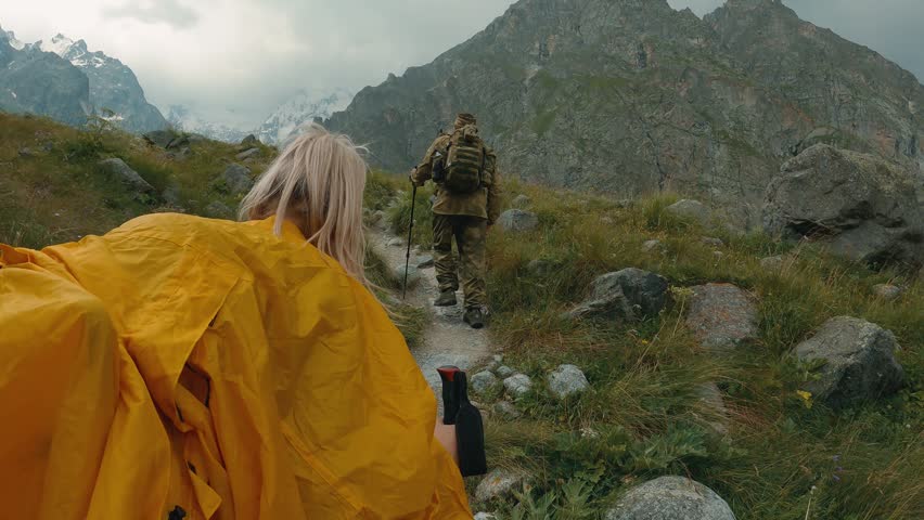 Two hikers walking uphill on rocky mountain path surrounded by green grass wildflowers. Snowy peaks in background under overcast sky. Yellow rain jacket trekking poles visible