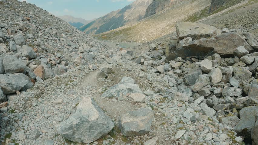 Mountain hiking path cuts through rocky terrain under bright sky. Gravel trail leads toward distant alpine lake rugged peaks. The camera moves over the path