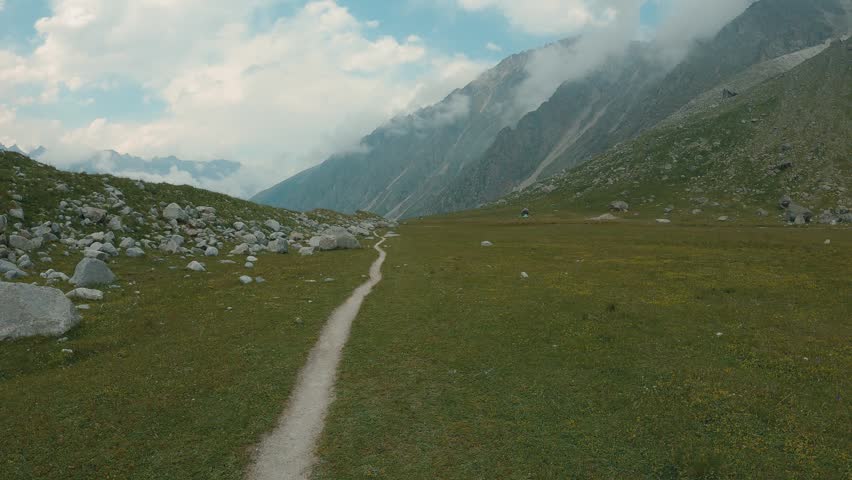 Winding dirt path cuts through vibrant green meadow dotted with wildflowers scattered boulders. Towering rocky mountains rise under soft clouds. The camera moves over the path