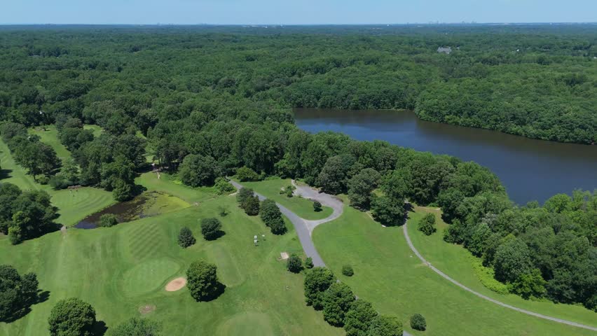 High-angle aerial drone footage capturing a beautiful golf course nestled in a dense, lush green forest. The scene features manicured fairways, a paved access road, and a tranquil lake bordering the property (Burke Lake, VA). The expansive woodland stretches to the horizon, highlighting a perfect summer day for outdoor recreation and sports. Ideal for projects showcasing country clubs, suburban parks, leisure activities, nature landscapes, and peaceful recreational real estate.