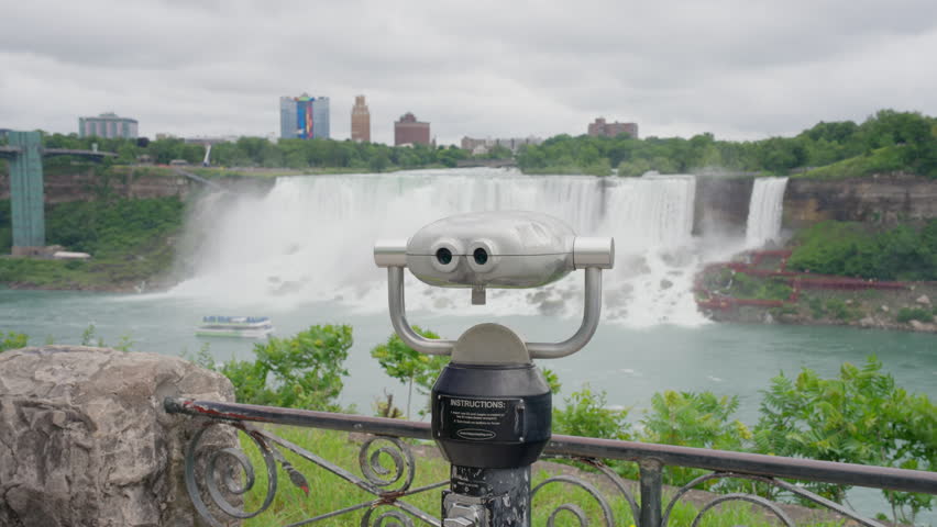 A coin-operated viewfinder stands before American Falls as a tour boat crosses the river and the U.S. skyline rises behind, Static Shot
