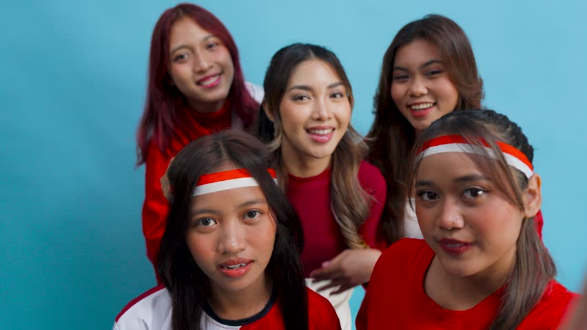 Five young Indonesian women is smiling happily while taking selfie together, isolated by blue background.