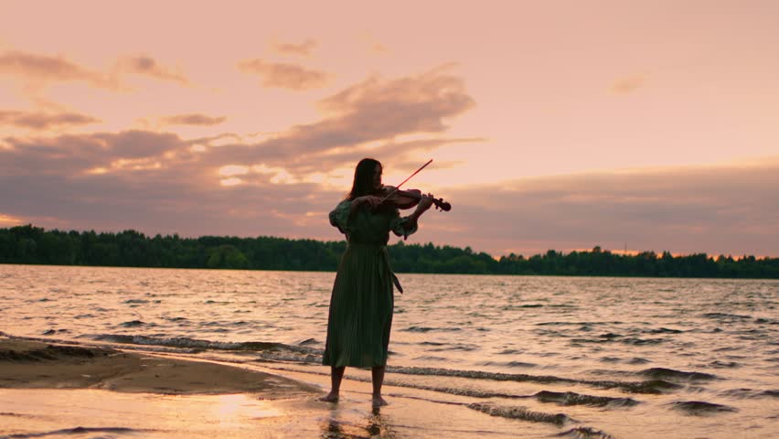 Woman playing violin on tranquil lakeshore at sunset, standing barefoot in gentle waves beneath dramatic cloudy sky, creating serene musical atmosphere in nature