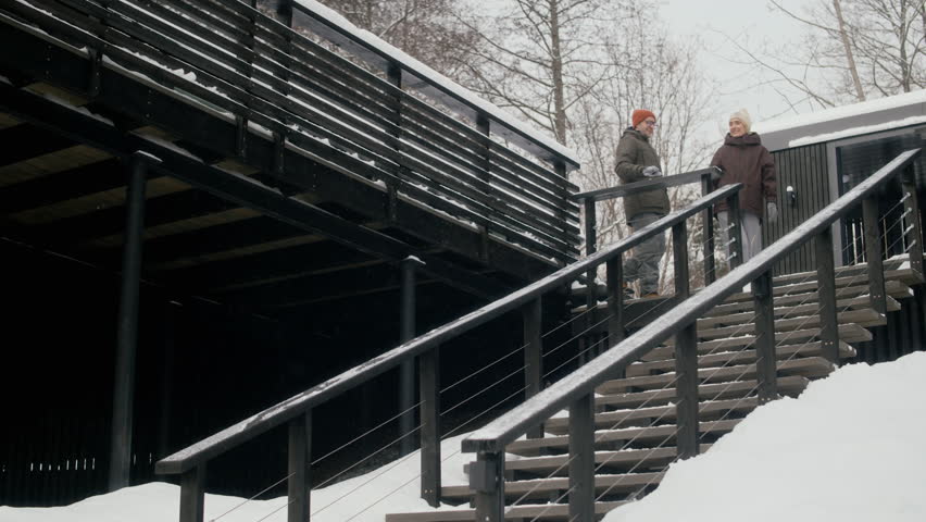 Caucasian adult couple walking down wooden stairs of modern cabin in winter, talking and smiling. Clip suitable for winter tourism ads and real estate marketing