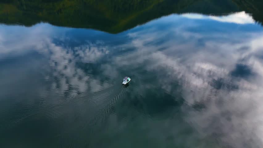 Aerial perspective of a small boat sailing on a beautiful, calm lake. The still water perfectly reflects the blue sky, clouds, and green hills