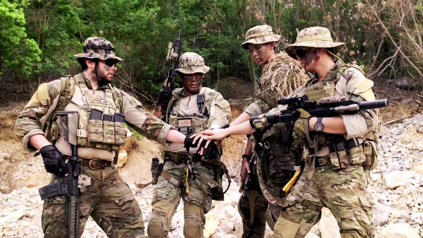 Four camouflaged soldiers in tactical gear form a team huddle outdoors, holding rifles during field training in a rugged, wooded area.