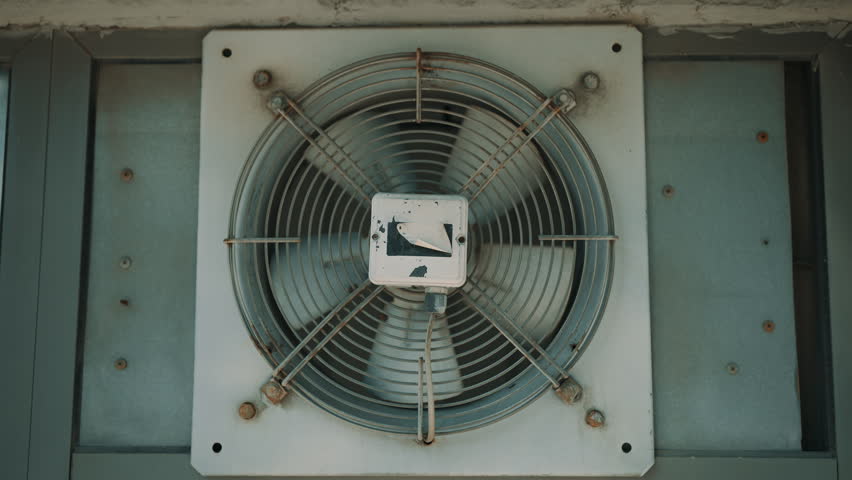 Weathered wall-mounted exhaust fan with protective grille, showing rust and wear. Industrial ventilation equipment close-up for airflow, cooling, and HVAC concepts.