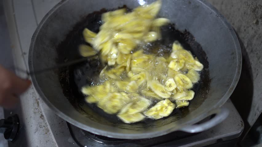 Close-up image of a woman traditionally and manually frying banana chips in a frying pan filled with boiling hot oil using a metal spatula.