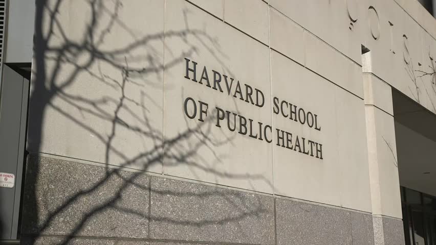 Close-up of the Harvard School of Public Health building sign in Boston, Massachusetts, with bare tree branches casting shadows across the facade.