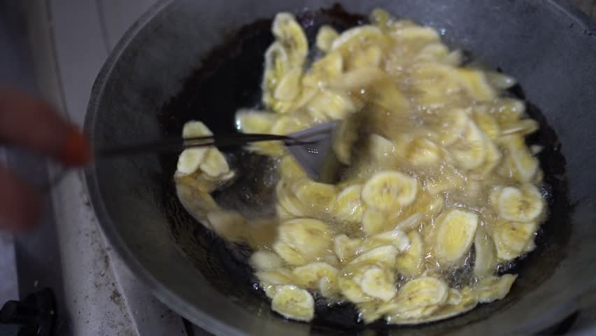 Close-up image of a woman traditionally and manually frying banana chips in a frying pan filled with boiling hot oil using a metal spatula.