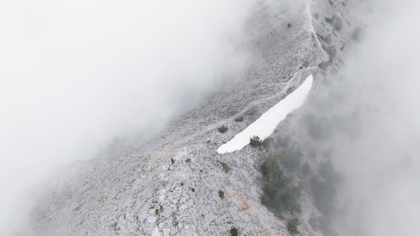 Atmospheric drone footage showing a caravan of pack mules ascending a rocky mountain ridge wrapped in thick fog and low clouds on Mount Olympus, Greece. Captures traditional supply transport, rugged alpine trails, and dramatic high-altitude weather conditions. Perfect for travel, documentary, culture, and outdoor adventure projects.