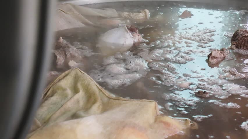 Close-up of spice pouches and meat simmering in a large pot of boiling broth with foam