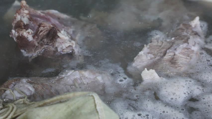 Close-up of meat and a spice pouch simmering in a large pot of boiling broth with foam