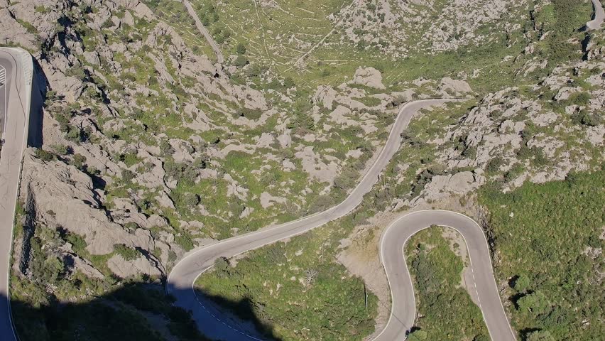 Aerial view shows a winding road cutting through rocky terrain in Mallorca, Spain. The scene captures the natural curves and greenery surrounding the road.