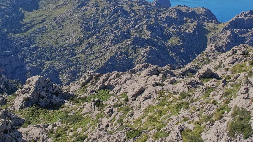 A view from above shows rocky terrain and greenery in Mallorca, Spain. The coastline can be seen in the distance. The area appears natural and untouched by buildings.