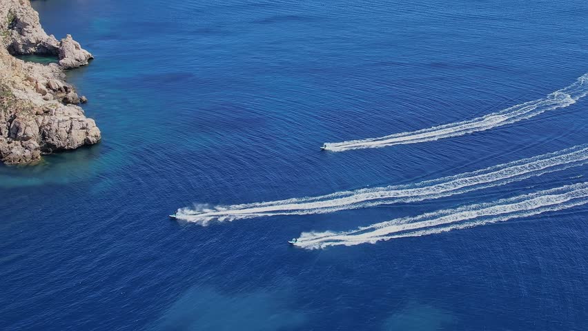 In Mallorca, Spain, several jet skis move quickly across the blue water near the rocky coastline. The sun shines brightly, highlighting the waves and splashes.