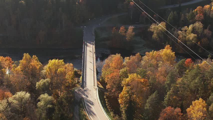 People walk across a bridge over a river in Sigulda. Surrounding trees show shades of yellow and orange as autumn sets in the area. A clear sky is visible above them.