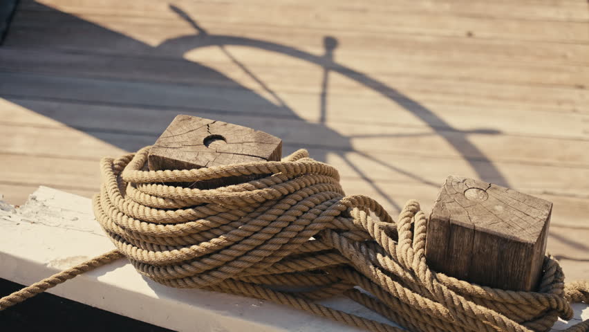 Close-up of a coiled mooring rope on weathered wooden posts with a ship wheel shadow in warm sunlight, featuring nautical textures, strong composition and copy space for maritime concepts.
