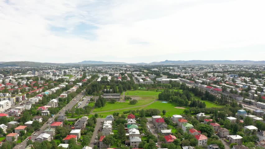 Aerial view of Reykjavik, Iceland, showing colorful residential houses, a large green park, and the cityscape with distant mountains under a cloudy sky.