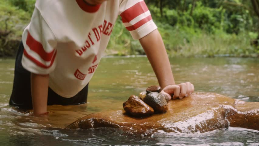 Close-up video of a child collecting and carefully stacking smooth river stones in a shallow forest stream, encouraging outdoor play, balance, creativity, and connection with nature.