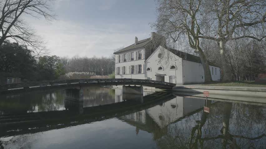 A wooden footbridge crosses a calm, reflective pond beside a white cottage in a quiet Parisian park in winter.