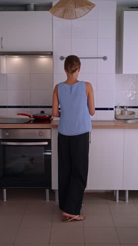 A person stands in a home kitchen, preparing food at the stove. The kitchen features white cabinets and a clean workspace. Morning light fills the room.