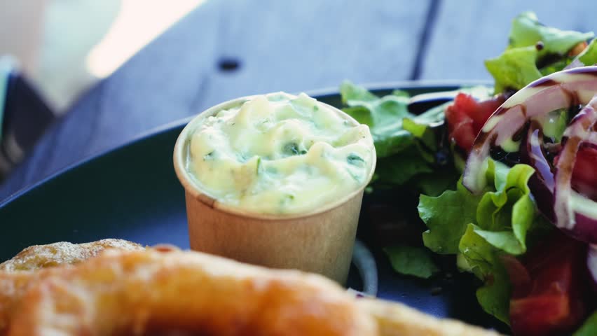 Close up of dipping a crispy onion ring into creamy sauce with fresh salad on the side. Appetizer snack and restaurant dining concept. High quality 4k footage
