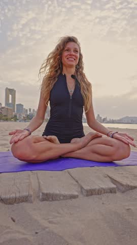 A woman performs yoga on a beach in Benidorm during early morning hours. The ocean and city skyline are in the background as she stretches and breathes deeply.
