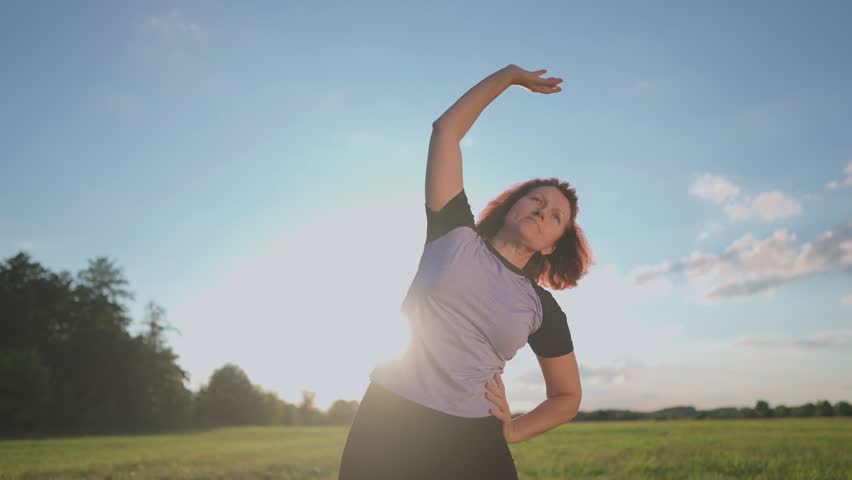 A woman practices sport by stretching her body outdoors. The woman focuses on her movements against a backdrop of green fields and blue skies.