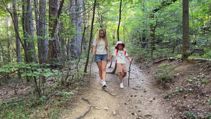 An adult woman and child approach along a forest path using wooden walking sticks while the camera moves backward ahead of them.