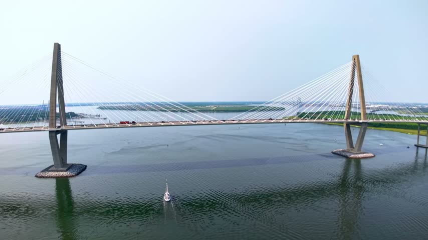 Aerial view captures Arthur Ravenel Jr Bridge over Cooper River during bright sunny day as sailboat glides underneath bridge structure while highway traffic flows across road deck above.