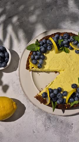 homemade lemon cheesecake topped with fresh blueberries, lemon slices and mint leaves, baked summer dessert under bright sunlight with harsh shadows