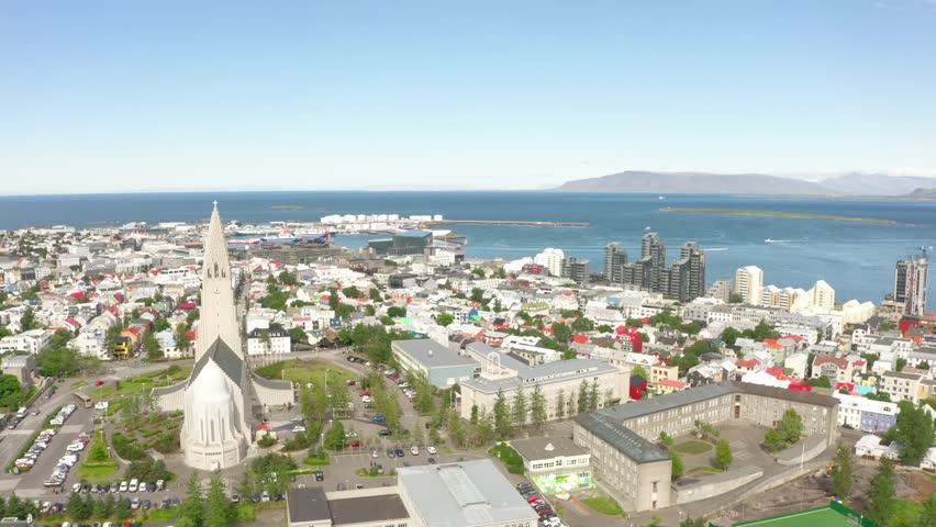 Wide aerial view of Reykjavik, Iceland on a sunny day. The iconic Hallgrímskirkja church is prominent in the cityscape, with the harbor and mountains in the background.
