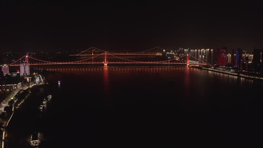 Wide night aerial shot of the red illuminated Yingwuzhou Yangtze River Suspension Bridge spanning across the Yangtze River in Wuhan, Hubei, China, framed by vibrant urban skyline lights.