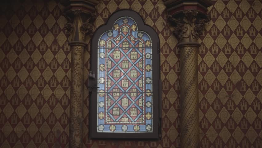 A small diamond patterned stained glass window glows softly in a dim, richly decorated chapel interior in Paris.