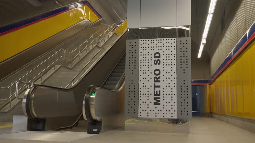 A tracking shot moving past a Metro SD branded pillar toward ascending escalators and concrete stairs inside a modern subway station