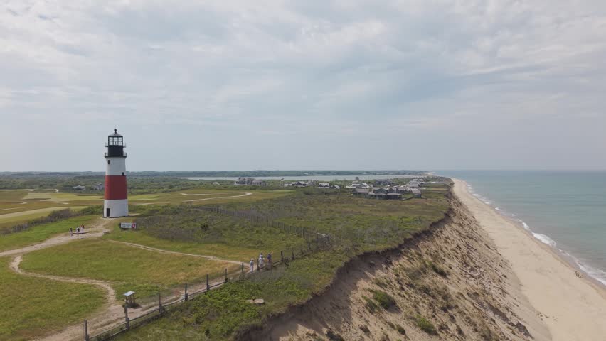 Sankaty Head Lighthouse stands red and white atop grassy bluff, visitors walk fenced trail along eroding sand cliff above sandy ocean beach in Nantucket, Massachusetts. Aerial drone wide shot