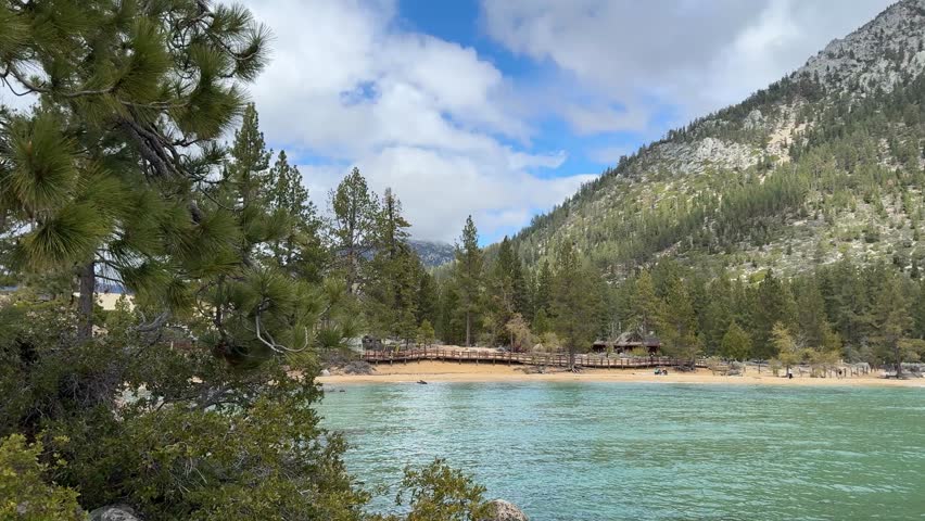Turquoise waters of Sand Harbor Beach at Lake Tahoe stretch toward a sandy shoreline backed by tall pine trees and rugged granite mountains under partly cloudy skies shifting to overcast