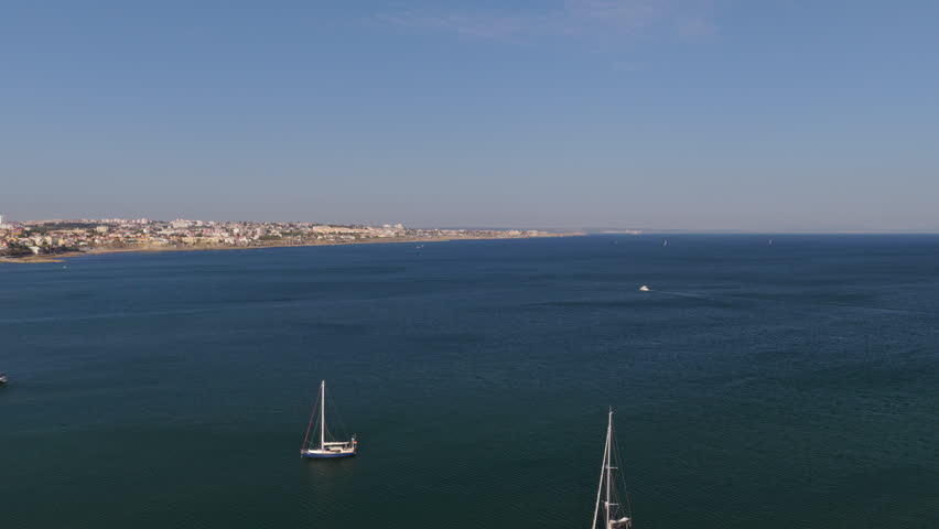 Wide drone panorama of Cascais Bay with anchored sailboats, the Atlantic Ocean and the Portuguese coastline stretching toward Estoril during golden hour.