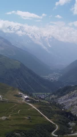 Vertical aerial drone shot moving slowly forward over an alpine valley near Mont Blanc in the French Alps, showing mountain trails, green slopes, distant village, cloudy peaks and glaciers in summer.