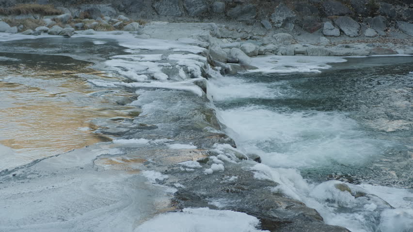 River waterfall frozen ice rocks, static handheld shot. Close up of small alpine stream cascade flowing through cold stones covered in winter glaze.