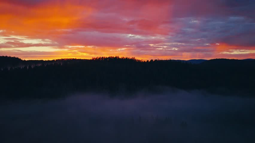 Aerial view of a sunset over a forested valley with mist drifting through the trees, layered hills, and warm evening light across the natural landscape