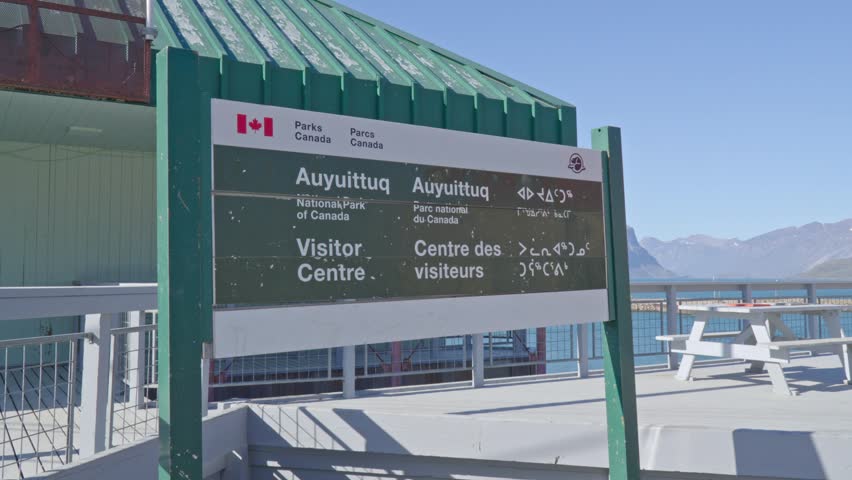 Ground-level shot of the official Parks Canada "Auyuittuq National Park Visitor Centre" sign framed against mountains and coastal fjord waters.