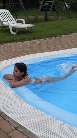 Young girl swimming and kicking along the edge of a small backyard pool on a sunny summer day, candid childhood outdoor fun and vacation leisure
